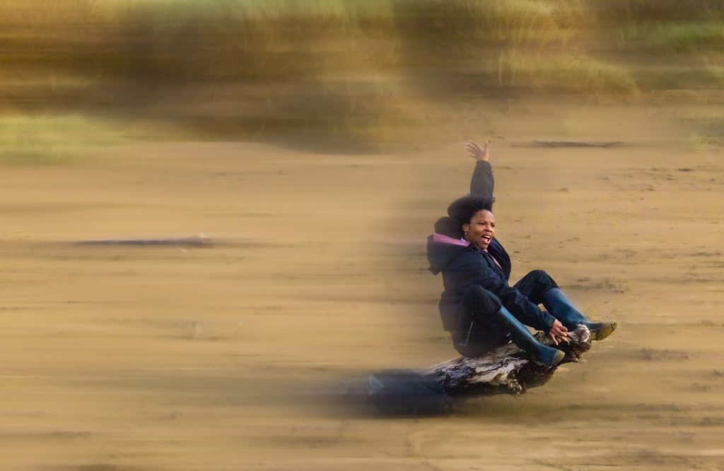 A Black woman is sat on a fallen tree trunk on a beach. She is leaning backwards with one hand holding onto the top of the trunk and one hand in the air. The trunk appears to be moving across the beach.